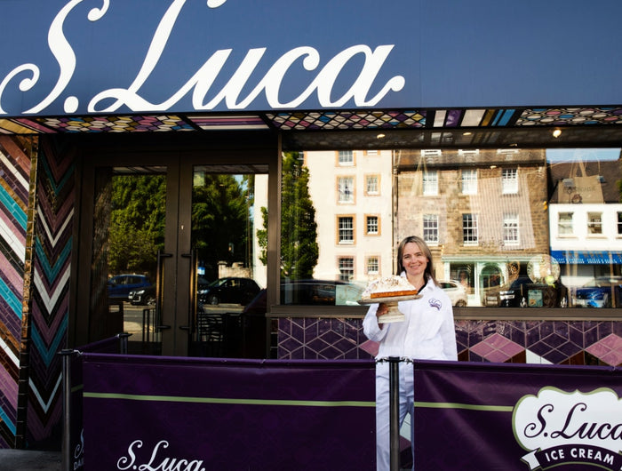 A woman in a white uniform stands in front of the S. Luca Ice Cream shop entrance, holding a cake.